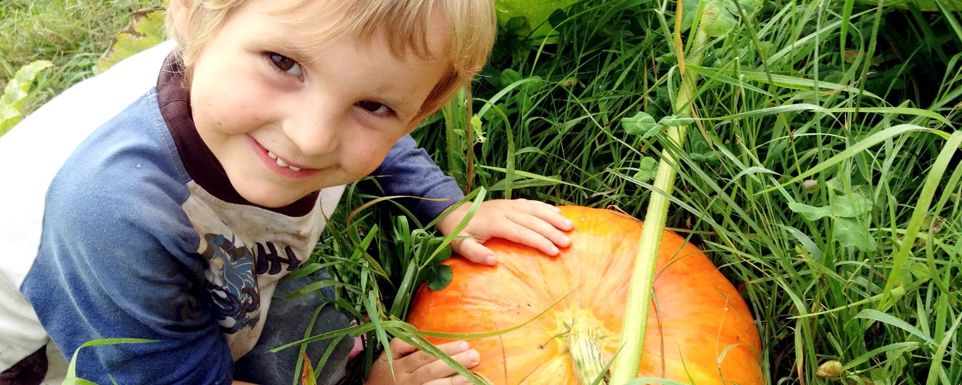 Un jeune garçon souriant pose les mains sur une grosse citrouille dans l’herbe